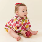 A baby wearing a white long-sleeve romper with a colorful fruit print, sitting on a cream background and holding a red apple.