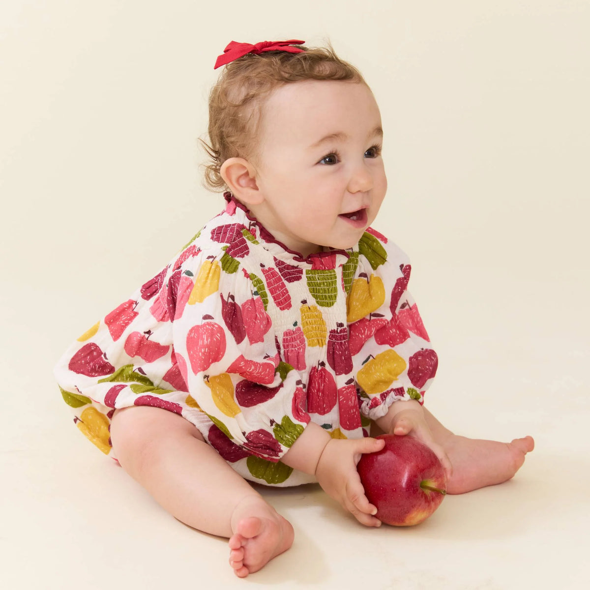 A baby wearing a white long-sleeve romper with a colorful fruit print, sitting on a cream background and holding a red apple.