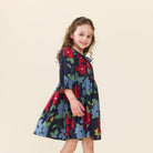 A young girl with curly brown hair wearing a navy blue dress with a colorful floral print, standing against a solid light beige background.