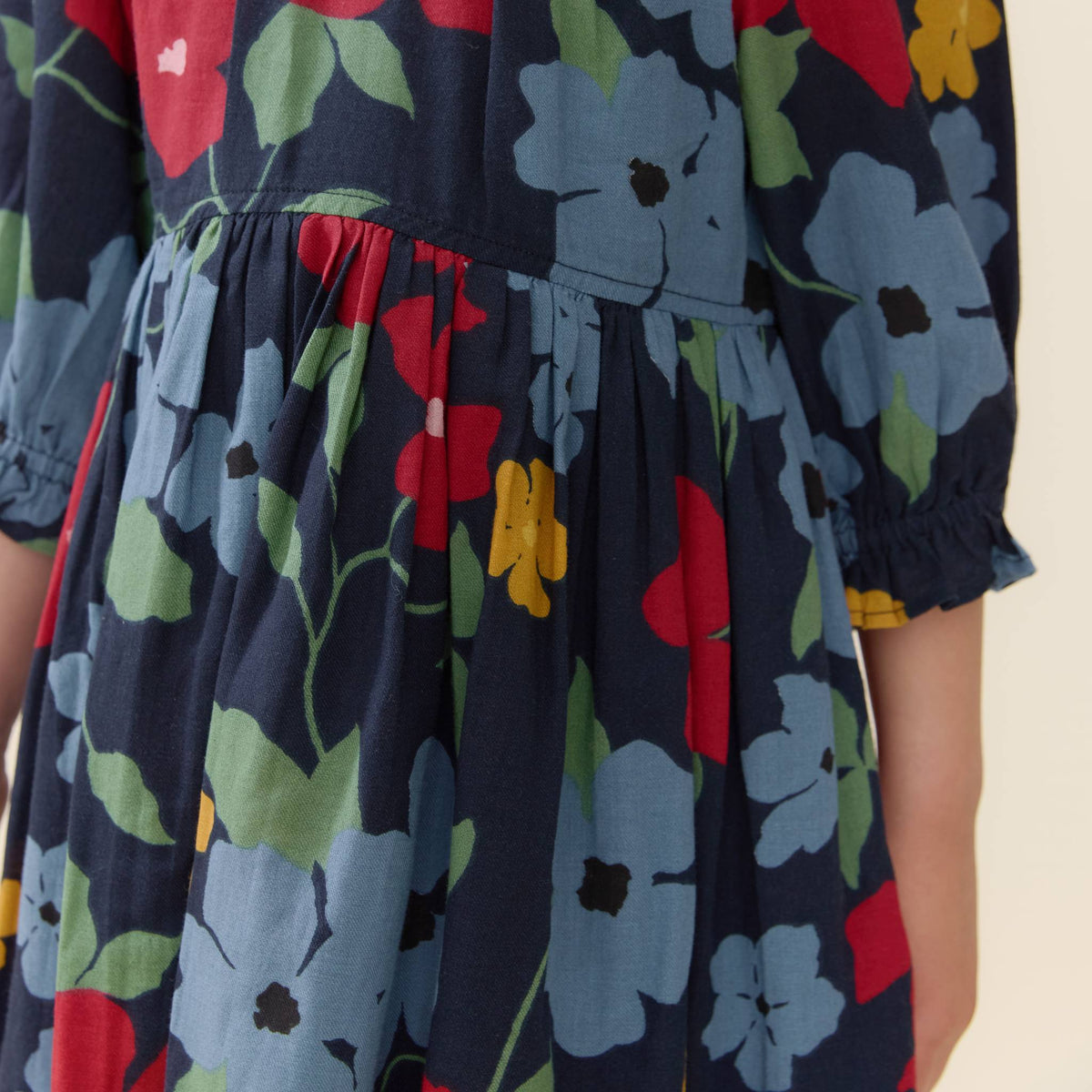 Close-up of a child wearing a navy blue dress with a colorful floral print in red, light blue, green, and yellow, featuring a gathered waist and ruffled sleeves.