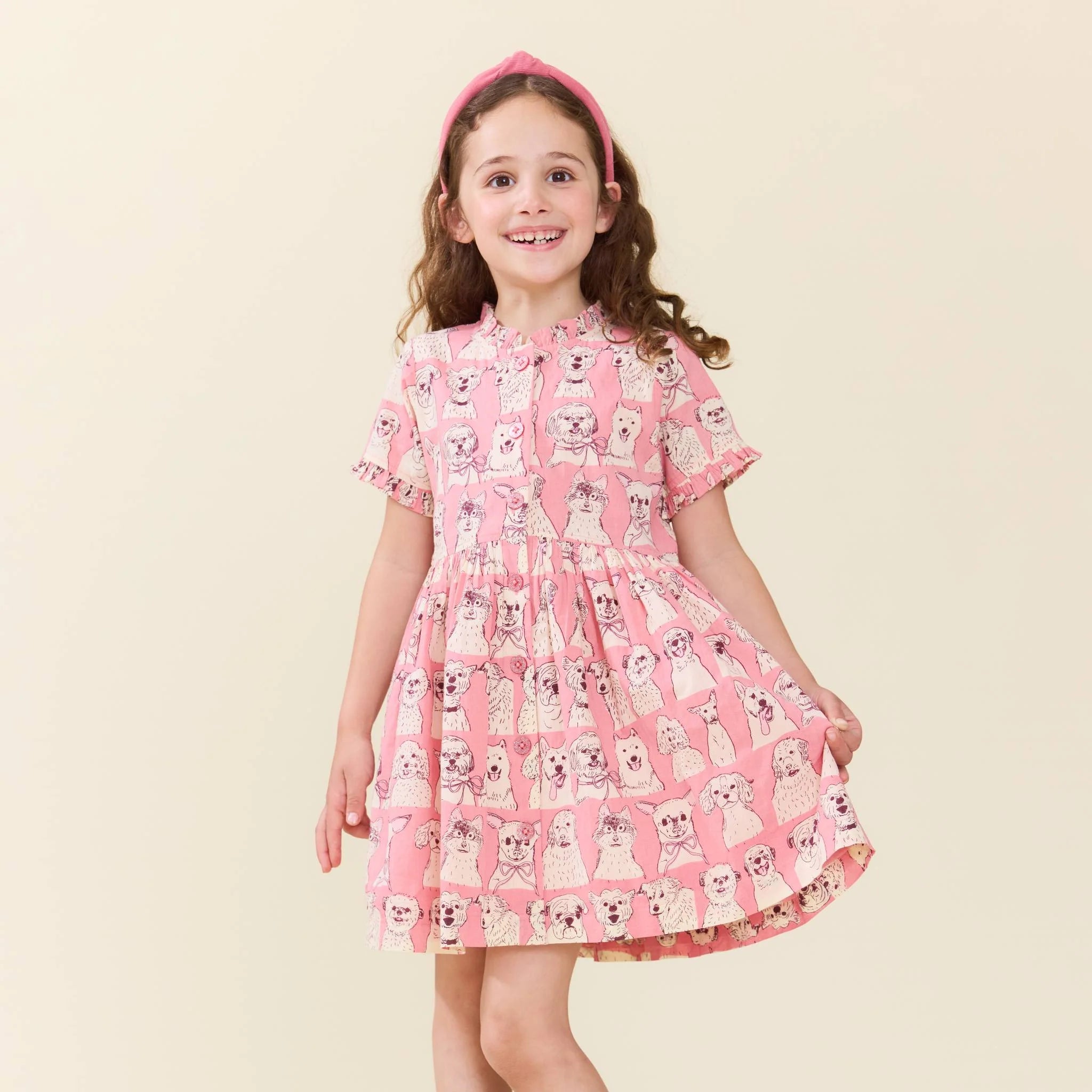 A young girl with long brown hair wearing a pink short-sleeve dress with a white and black dog pattern, a pink headband, and smiling against a solid light beige background.