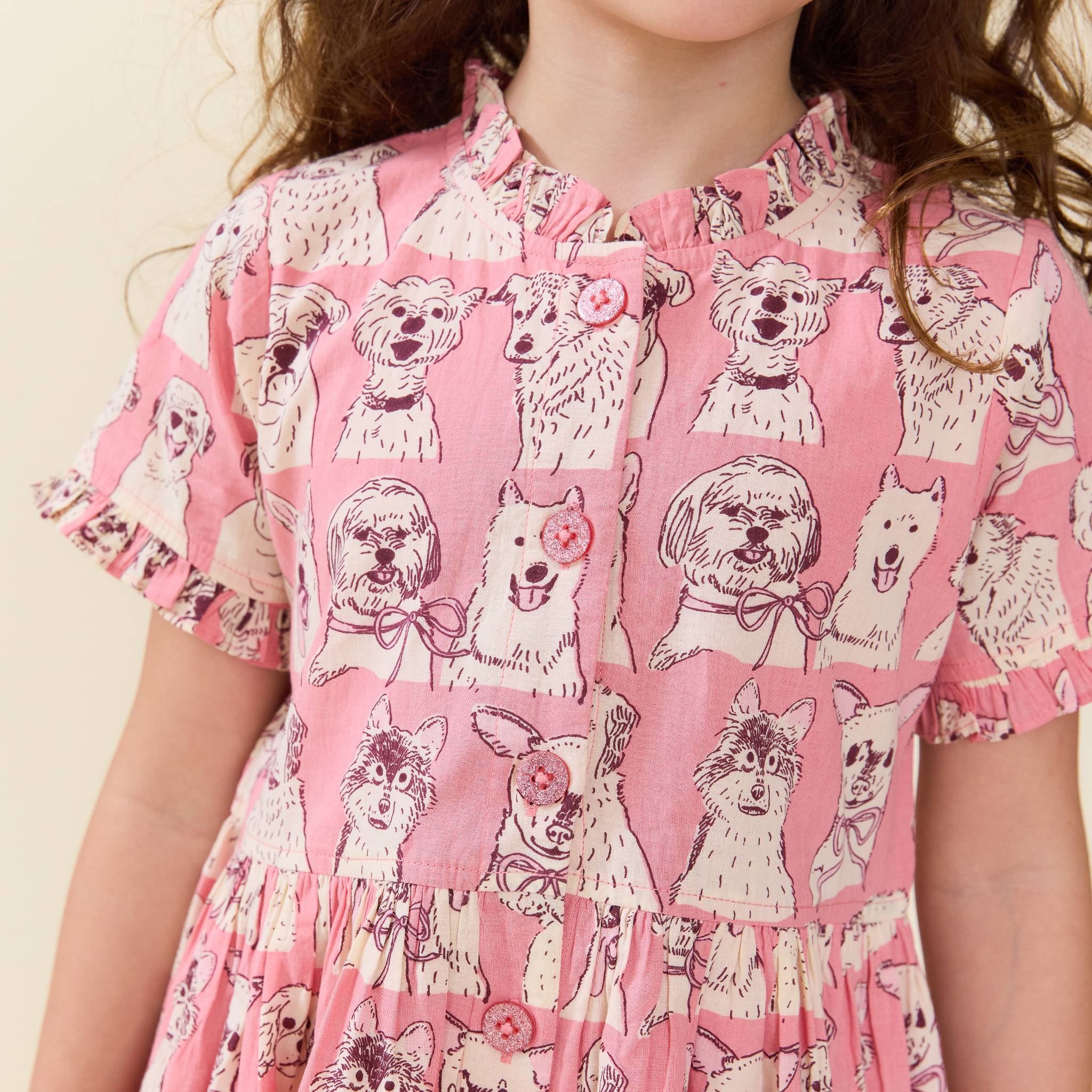 A close-up of a child wearing a pink short-sleeve dress with an allover print of different dog illustrations in cream and dark pink. The dress features a ruffled collar, a buttoned front, and ruffled sleeves.