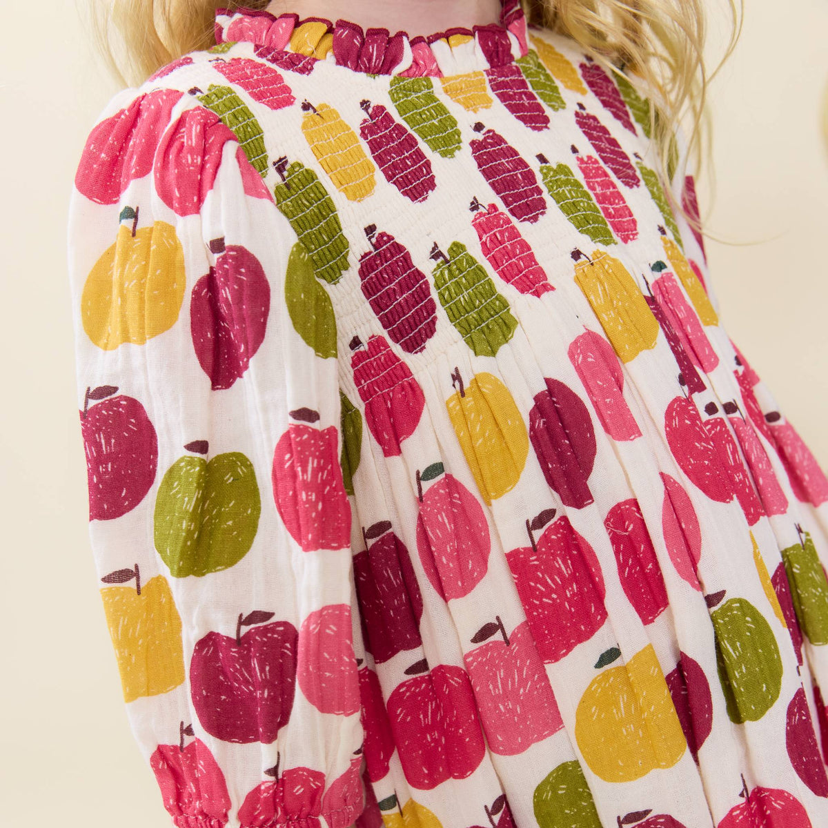 A close-up of a child wearing a white long-sleeve blouse with a colorful fruit print featuring pink apples, yellow pears, and green lemons. The blouse has a ruffled neckline and is shown against a light beige background.