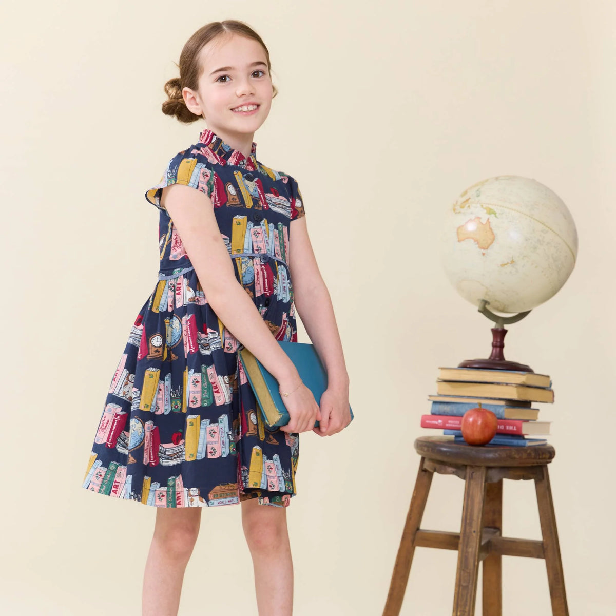 A young girl stands in a studio wearing a navy blue dress with a colorful book-print pattern. She is holding a blue book and standing next to a wooden stool with a vintage globe and books on it, against a solid beige background.