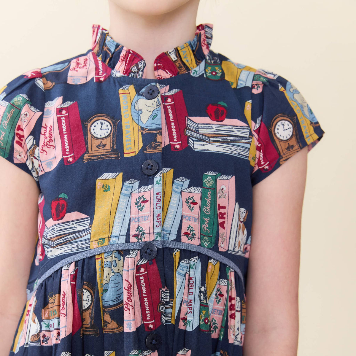 A close-up of a child wearing a navy blue dress with a colorful book-and-object print, featuring short cap sleeves, a ruffled collar, and front buttons, against a plain light beige background.