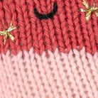 Close-up of a knitted textile with a coral-red upper section, a pale pink lower section, a small black curved detail near the top center, and small shiny gold bow accents.