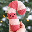 A hand holds a knitted candy cane plush toy with pink and white stripes, embroidered facial features, and a small green leaf and pink flower accent, against a blurred Christmas tree background with warm lights.