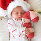 A sleeping newborn baby wearing a white waffle-knit onesie with red candy cane patterns and a red Santa hat, holding a red and pink knitted candy cane plush toy.