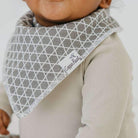 Baby wearing a gray patterned bib with a brand label, on a plain background