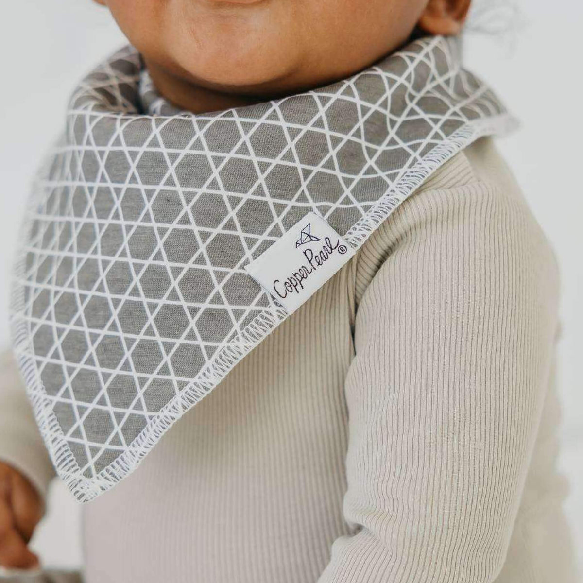 Baby wearing a gray patterned bib with a brand label, on a plain background