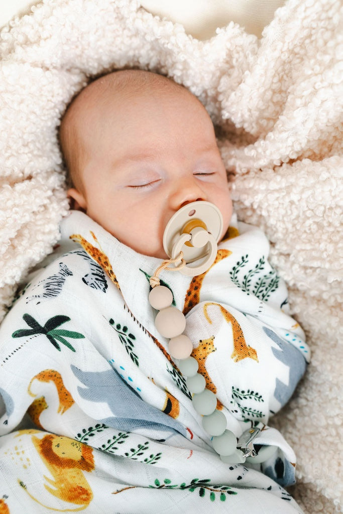 A sleeping newborn baby swaddled in a white blanket with a safari animal print, lying on a soft cream fleece blanket. The baby has a pacifier with wooden beads attached.