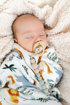 A sleeping newborn baby swaddled in a white blanket with a safari animal print, lying on a soft cream fleece blanket. The baby has a pacifier with wooden beads attached.