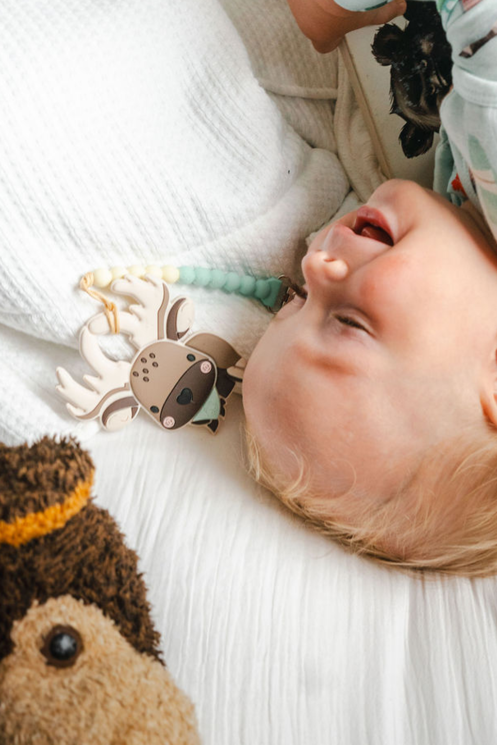 A close-up lifestyle shot of a baby lying on a white textured blanket, wearing a light blue patterned outfit and interacting with a wooden moose teething toy. A brown plush toy is visible in the foreground.