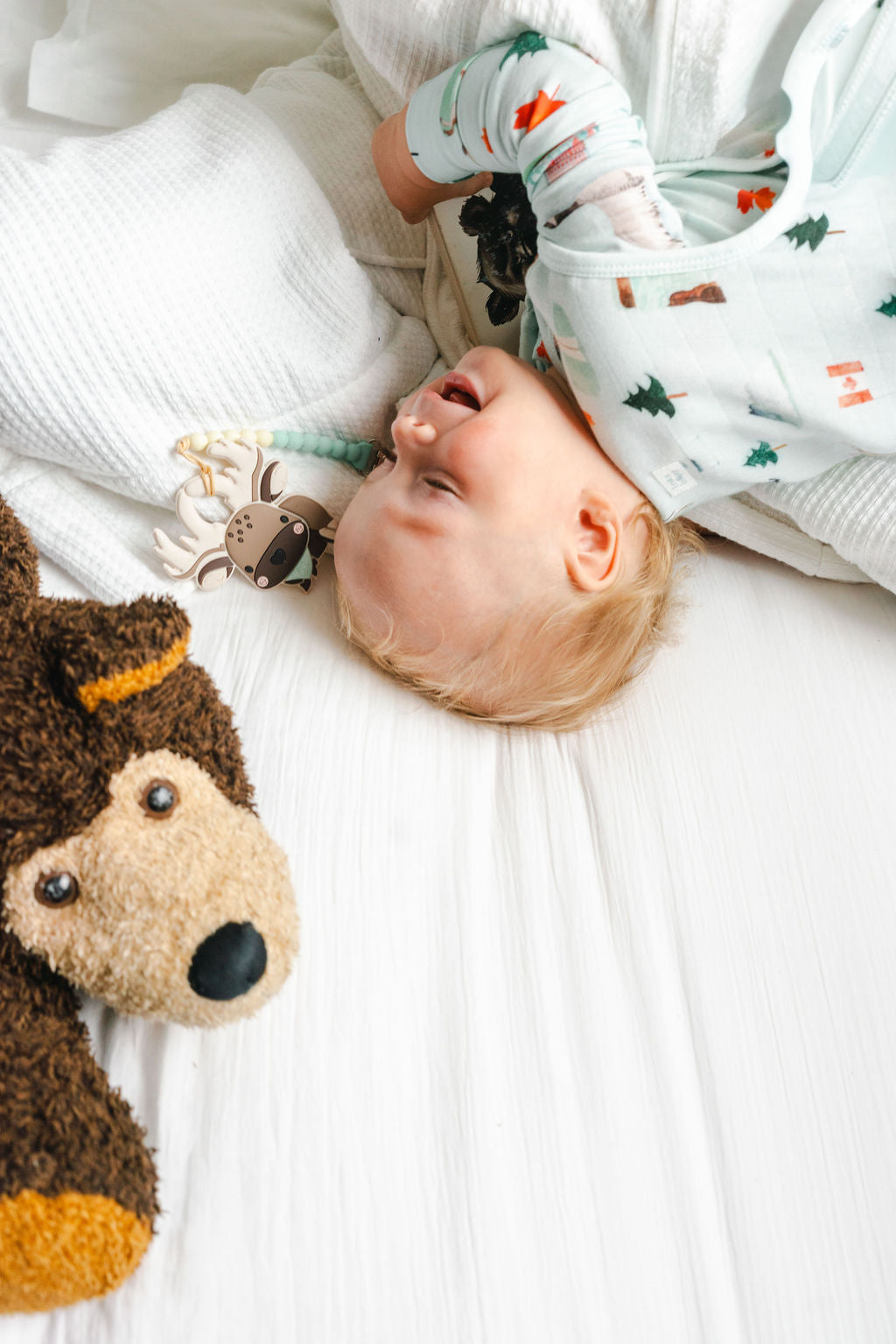 A baby lying on a white textured blanket, wearing a light green long-sleeve onesie with a forest pattern. A brown plush toy is visible in the foreground.