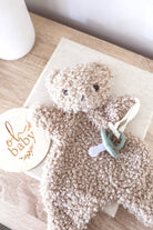 A oatmeal-colored, curly teddy bear plush toy with small black embroidered eyes and nose on a white background.