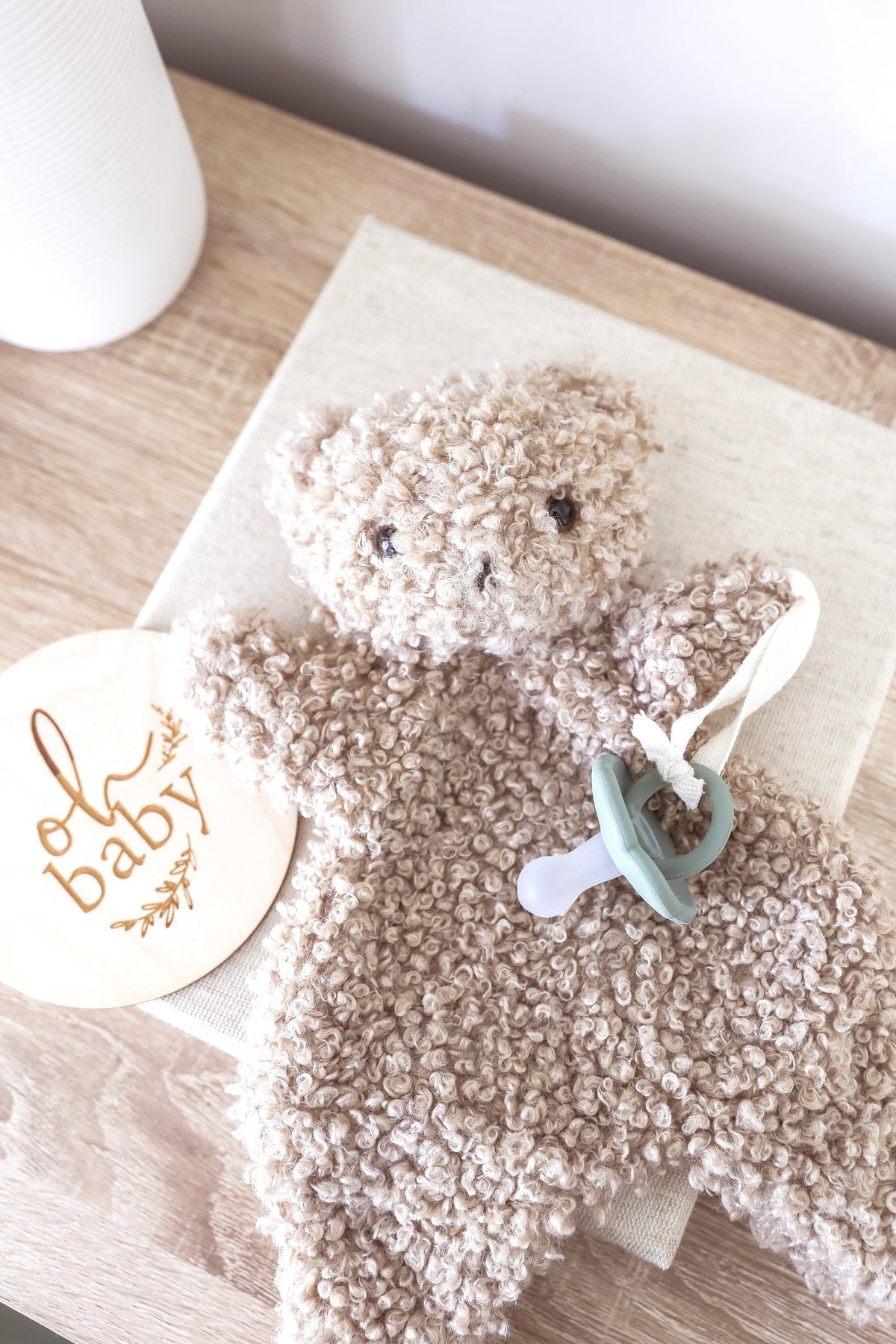 A oatmeal-colored, curly teddy bear plush toy with small black embroidered eyes and nose on a white background.