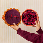 A person holds two round felt-like decorative pieces against a light beige striped background. One piece has a dark purple center with red and black ribbon crosses on an orange scalloped border.