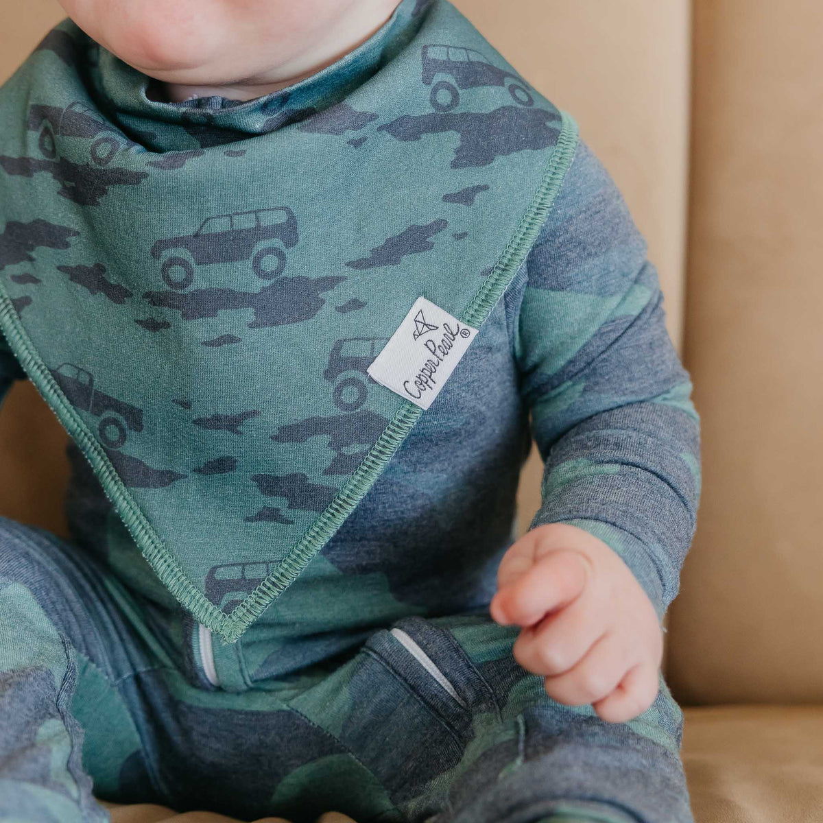 Child wearing a green bib with vehicle pattern and a branded tag, sitting on a beige surface.