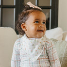 Baby wearing a floral bib and checkered outfit sitting on a couch.