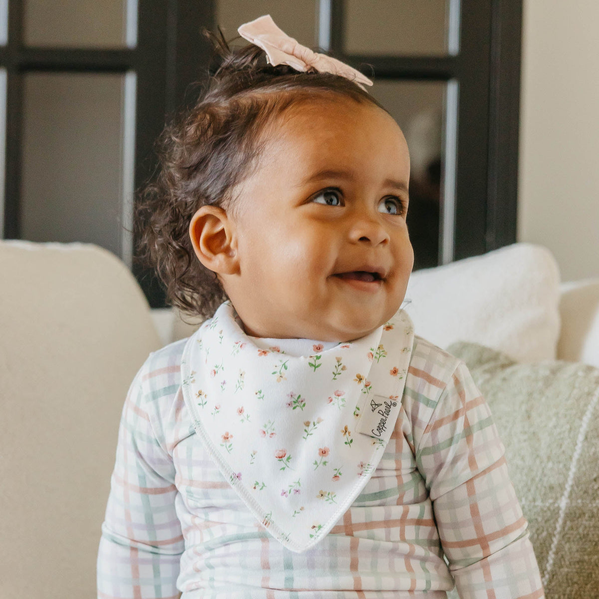Baby wearing a floral bib and checkered outfit sitting on a couch.