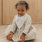 Child wearing a light-colored dress with a matching bib, sitting on a wooden floor.