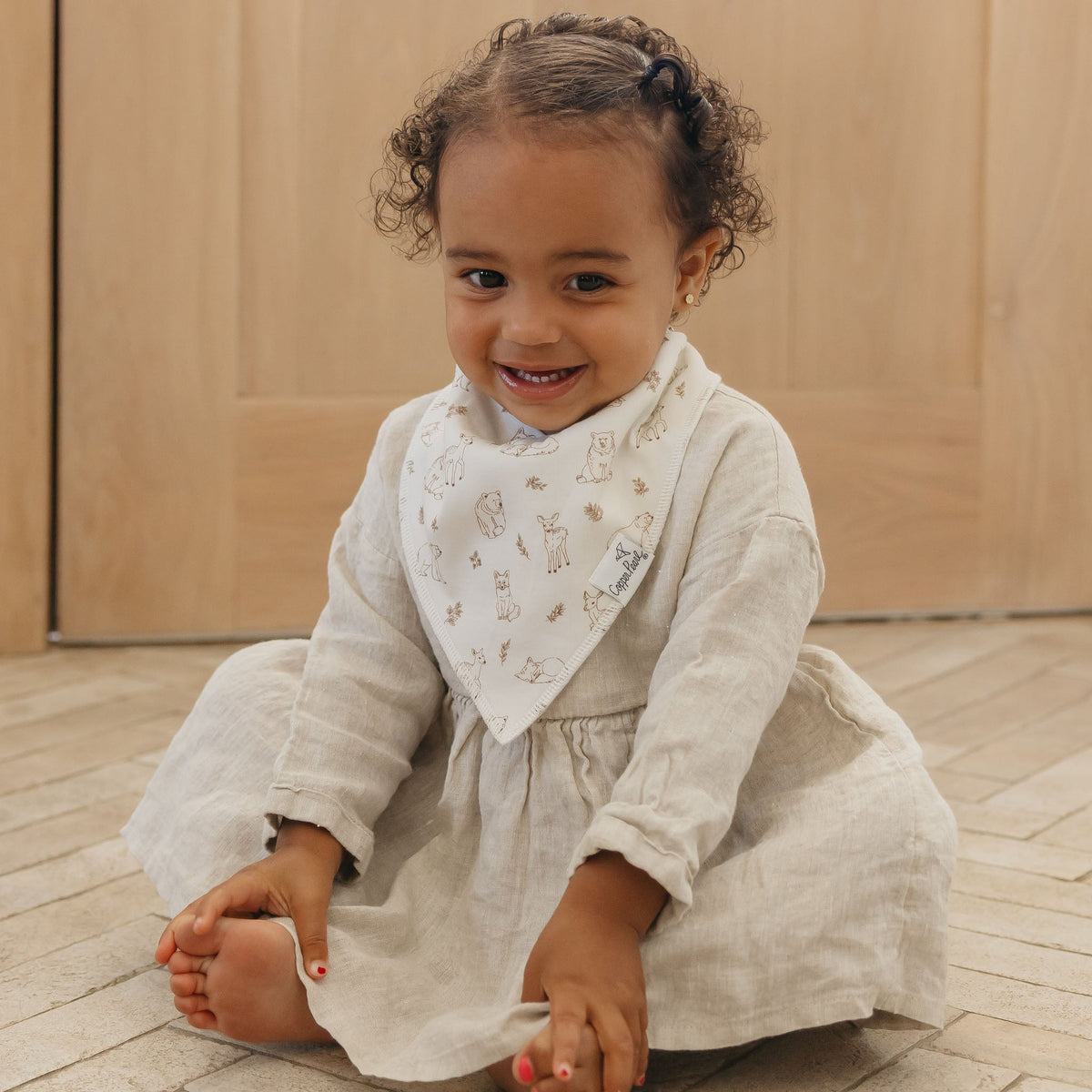 Child wearing a light-colored dress with a matching bib, sitting on a wooden floor.