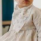 Child wearing a beige dress with a patterned bib and a visible brand label.