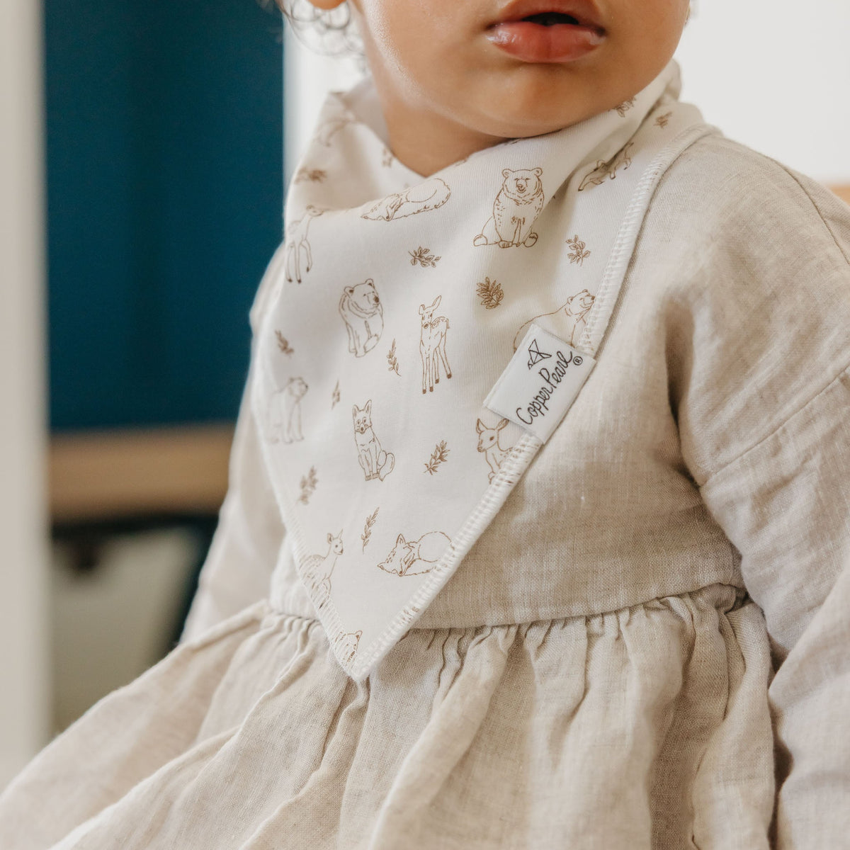 Child wearing a beige dress with a patterned bib and a visible brand label.