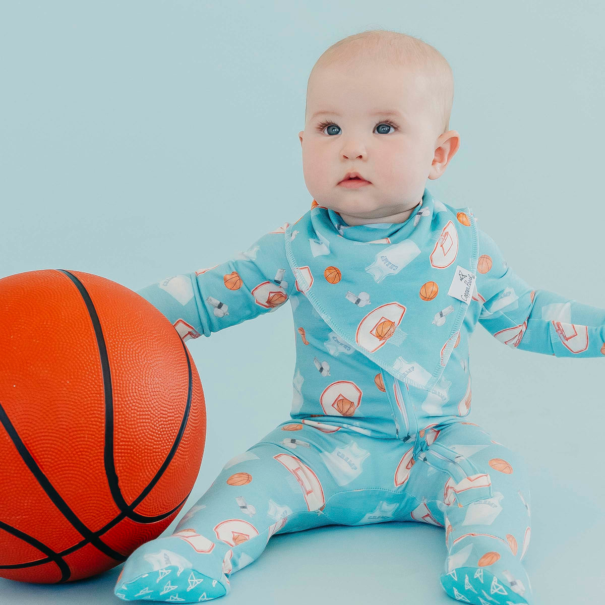 A baby sitting on a light blue studio background wearing a light blue footed sleeper with a basketball and whistle pattern, next to an orange basketball.