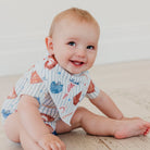 A smiling baby sitting on a light wooden floor wearing a short-sleeve bib with a white pinstripe pattern and colorful cartoon sea creature prints.