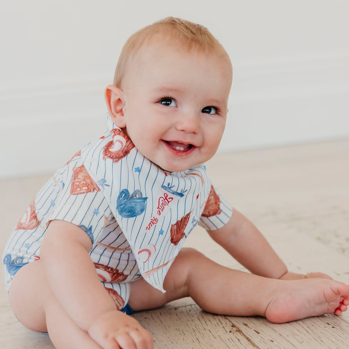 A smiling baby sitting on a light wooden floor wearing a short-sleeve bib with a white pinstripe pattern and colorful cartoon sea creature prints.