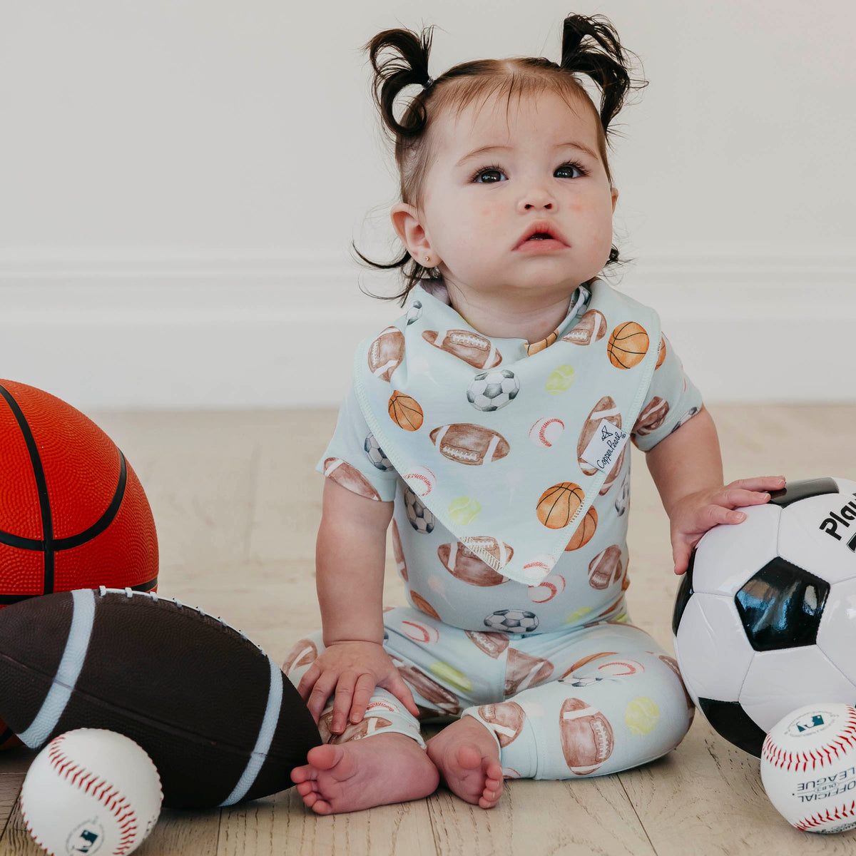 A baby girl with two pigtails sits on a light wood floor surrounded by sports balls, wearing a light blue short-sleeve pajama set with a football and basketball pattern.