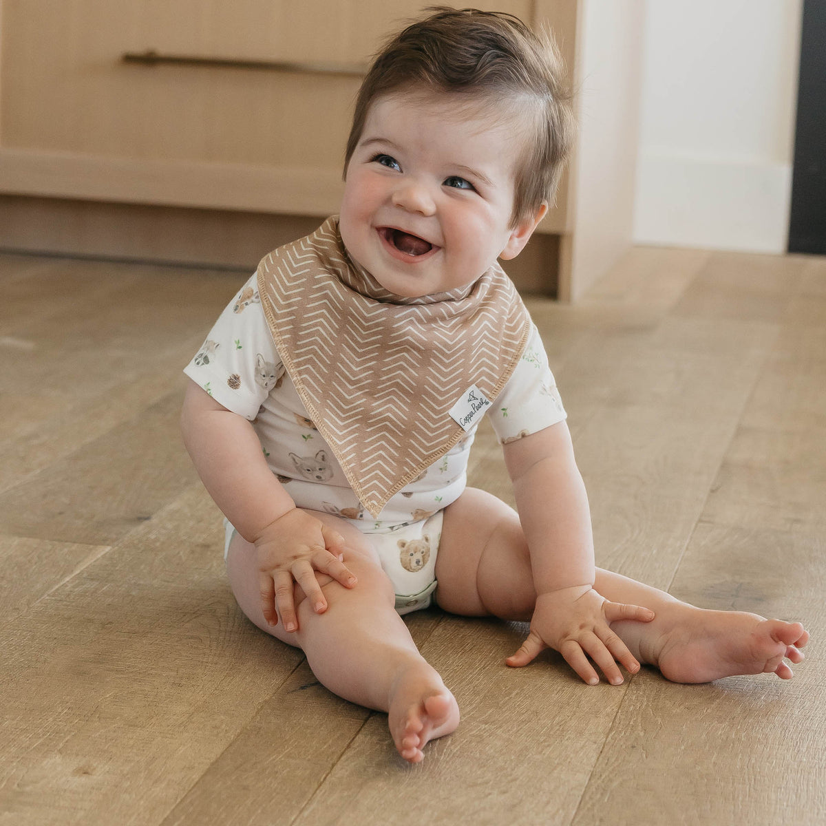 Baby sitting on a wooden floor wearing a bib and smiling.