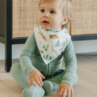 Baby wearing a green outfit with a forest-themed bib sitting on a wooden floor.