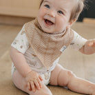 Baby wearing a beige bib with a pattern, sitting on a wooden floor.
