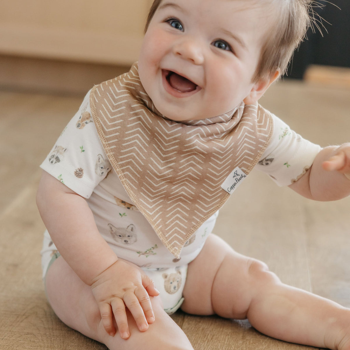 Baby wearing a beige bib with a pattern, sitting on a wooden floor.