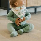 Baby in green outfit with forest-themed bib sitting on a wooden floor.