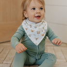 Baby in a green outfit with a patterned bib sitting on a wooden floor.