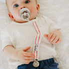 Baby wearing a white shirt with a red and white striped pacifier clip.