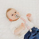 Baby lying on a white blanket wearing a white onesie with pink trim and blue pants.