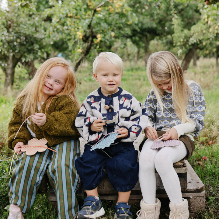 Three children sitting outdoors on a bench in a grassy, tree-filled setting, each holding and working on a small handmade paper craft.