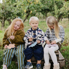 Three children sitting outdoors on a bench in a grassy, tree-filled setting, each holding and working on a small handmade paper craft.