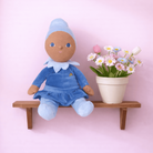 Doll in a blue outfit sitting on a wooden shelf with a pot of flowers against a pink background