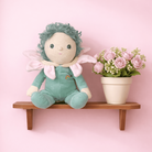 Stuffed fairy toy with green hair and outfit on a wooden shelf with a pink background