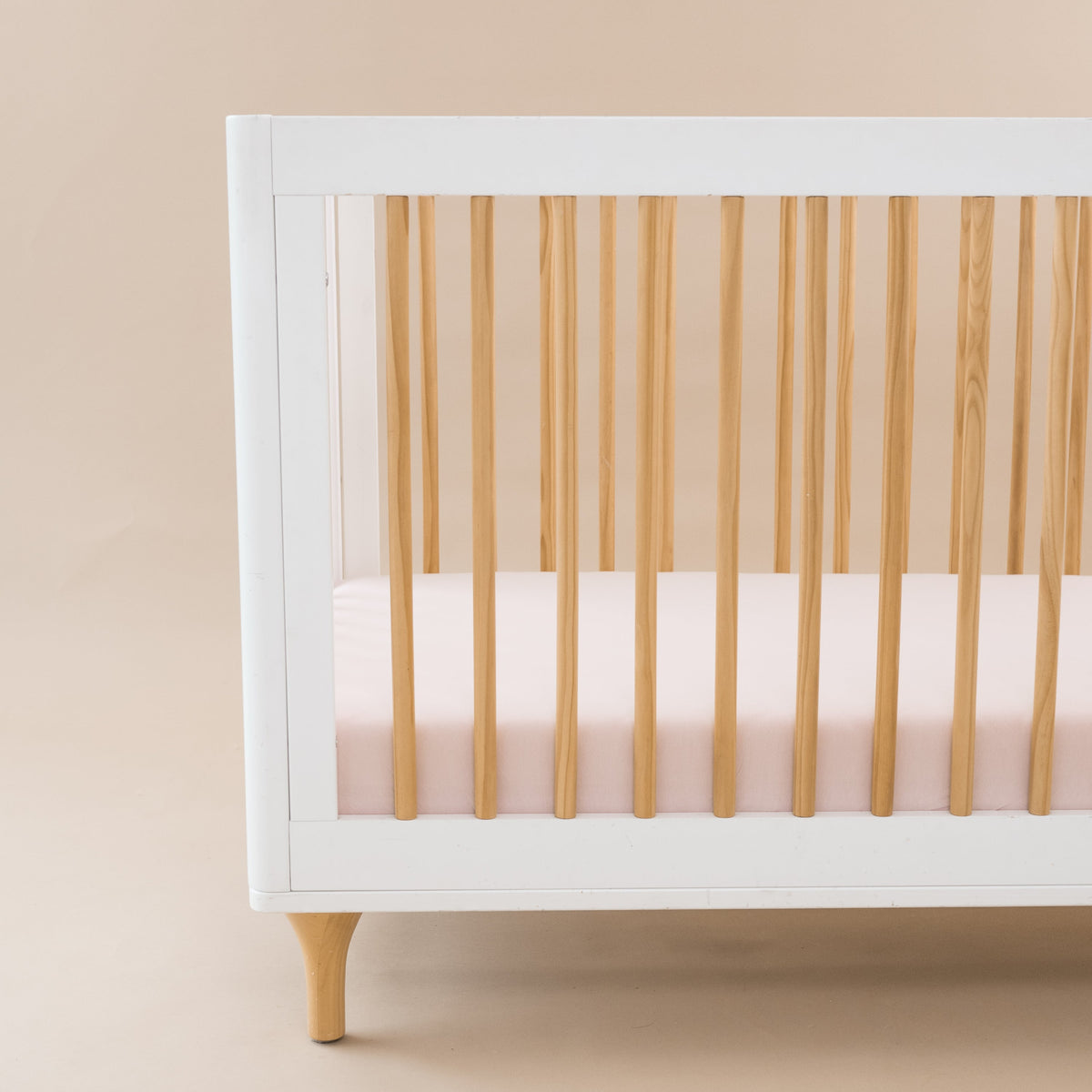 White crib with wooden sides and a pink mattress against a beige background