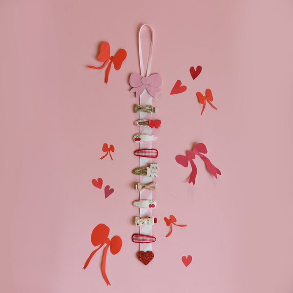 A vertical hair clip garland hanging on a pink background, decorated with multiple small hair clips, bows, and heart-shaped embellishments. Red and orange paper hearts and ribbon bows are scattered around the display.