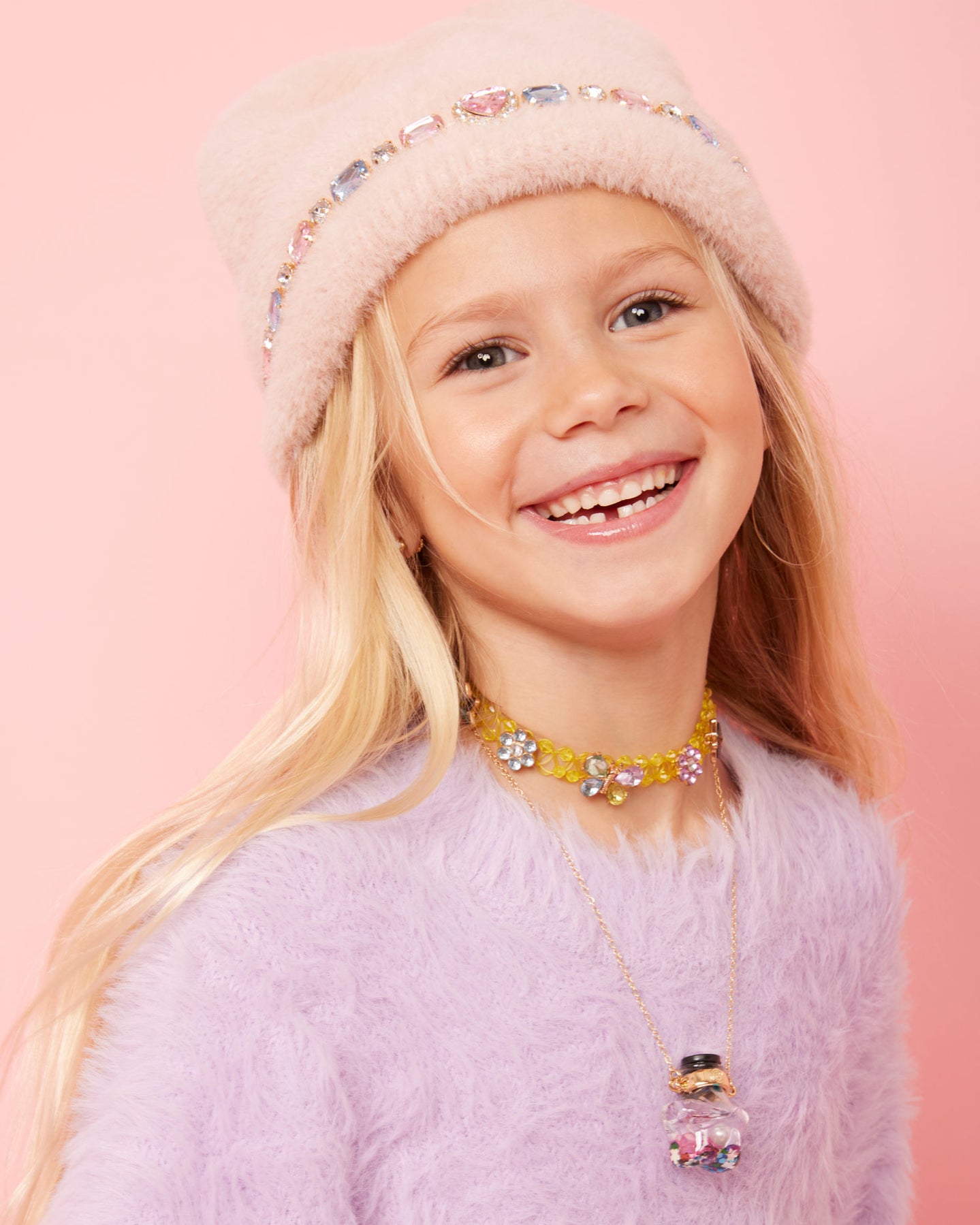 Young girl wearing a pink fluffy hat and purple sweater with a pink background