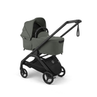 A stroller travel system in muted sage green with a black frame and wheels, shown on a clean white background. The system includes a bassinet attached to a stroller with a long handle and a black storage basket.