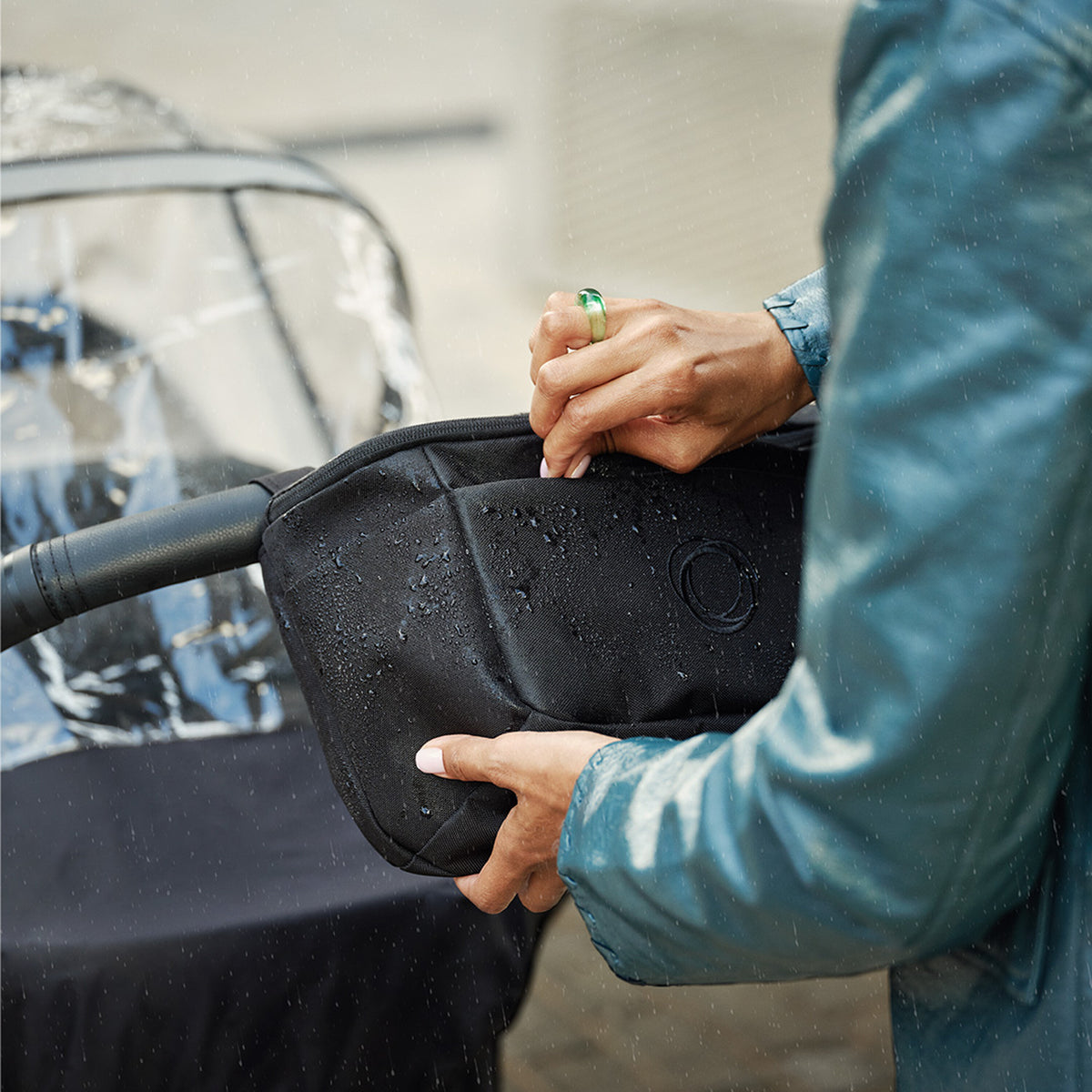 Person holding a black rain cover over a stroller with water droplets on it.