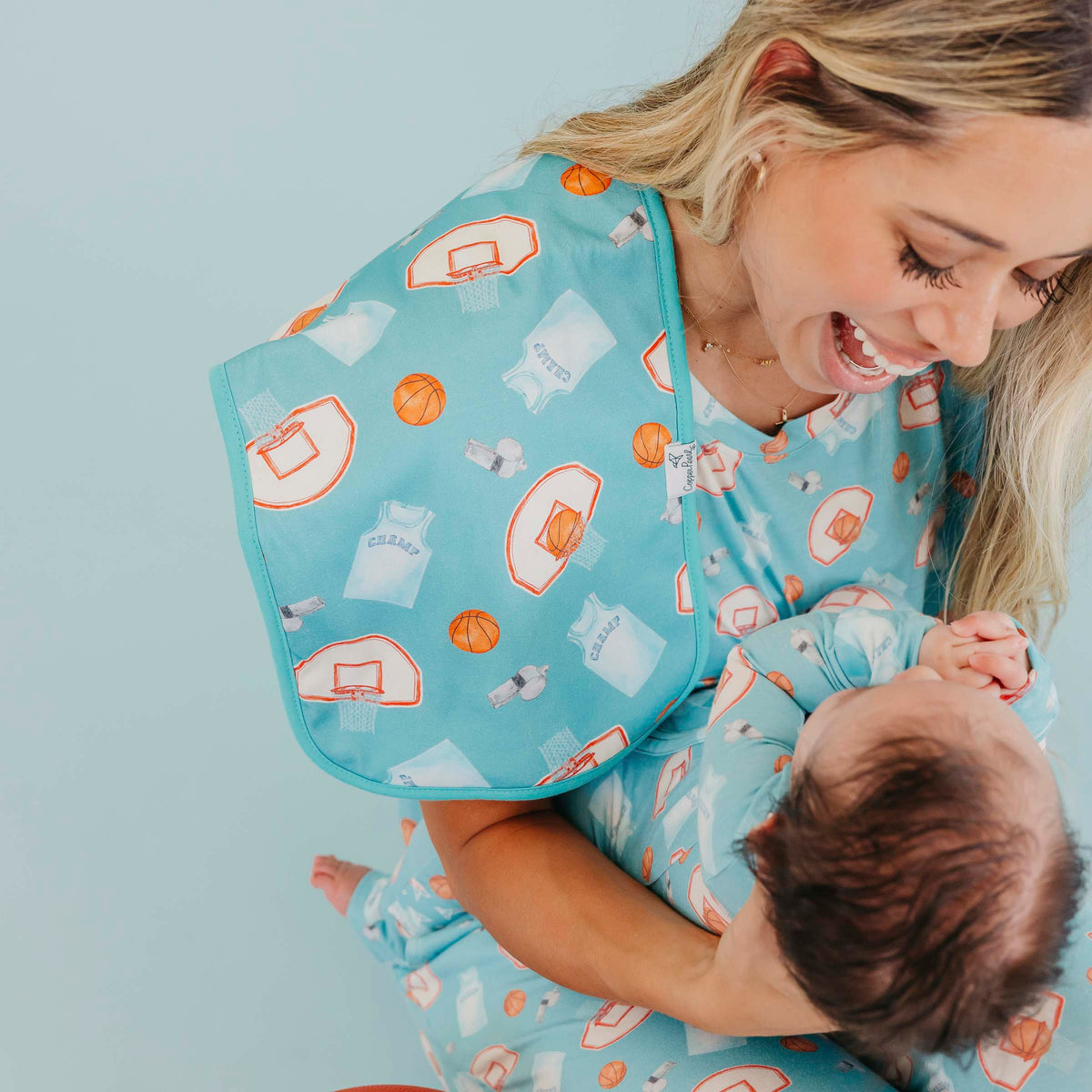 A smiling woman holds a baby wearing matching light blue printed pajamas and a bib with a basketball court and jersey pattern, against a pale blue background.